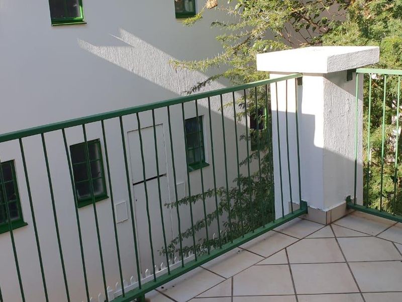A private balcony featuring tiled flooring and a green metal balustrade, overlooking the building exterior and surrounding greenery.