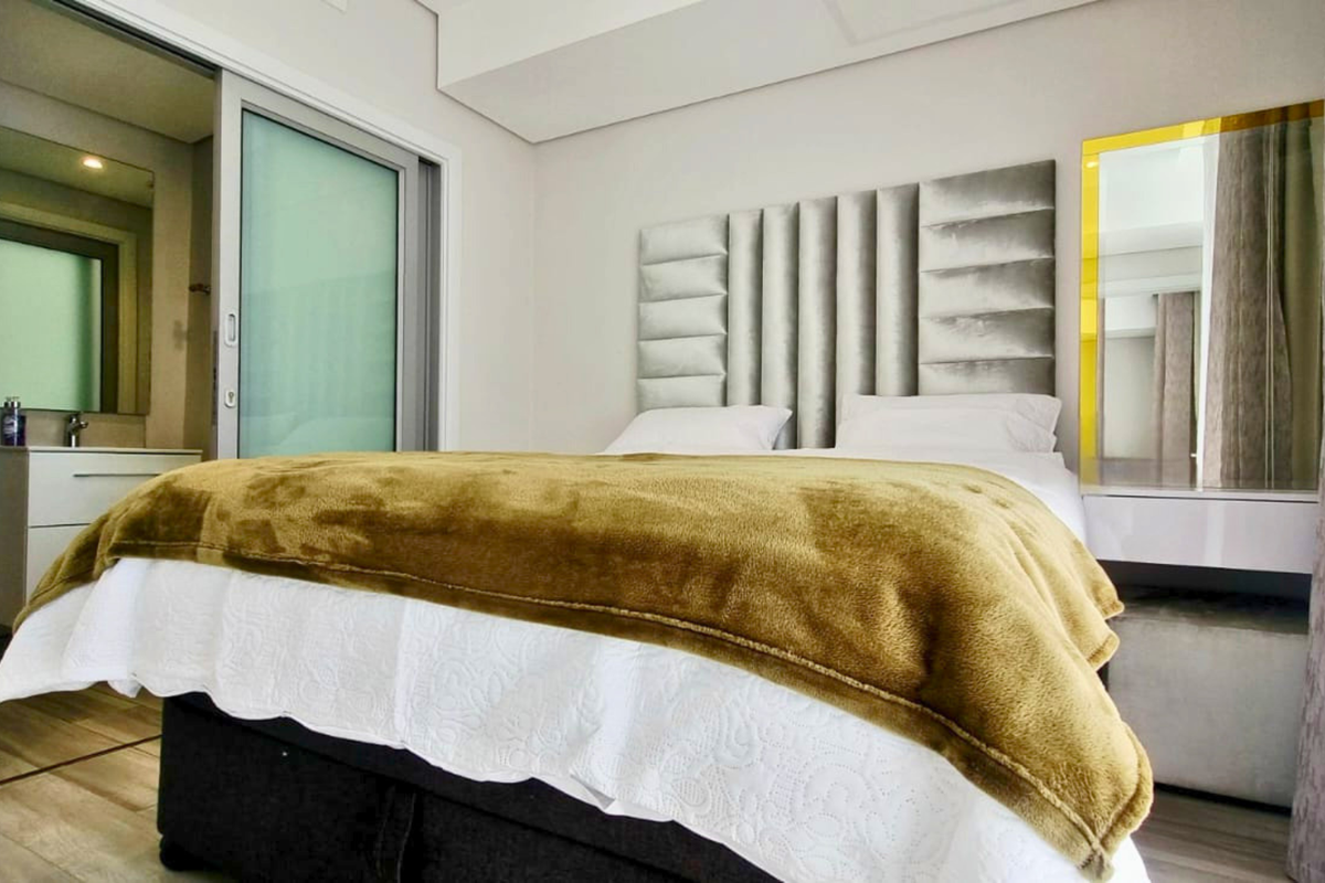 A modern bedroom featuring a built-in upholstered headboard and an en-suite bathroom accessible via sliding glass doors.