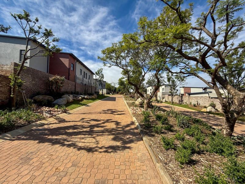 A well-maintained paved driveway within a residential cluster estate, featuring landscaped garden beds, mature trees, and brick boundary walls.