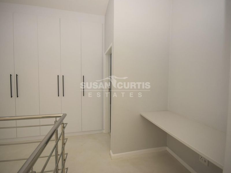 A clean, modern hallway featuring extensive built-in storage cupboards, a sleek stainless steel balustrade, and a convenient built-in desk area.
