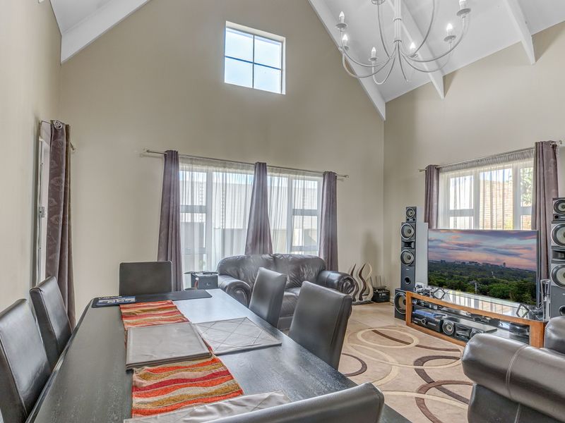 Spacious open-plan living and dining area featuring a high vaulted ceiling with exposed rafters, large windows for natural light, and a central chandelier.
