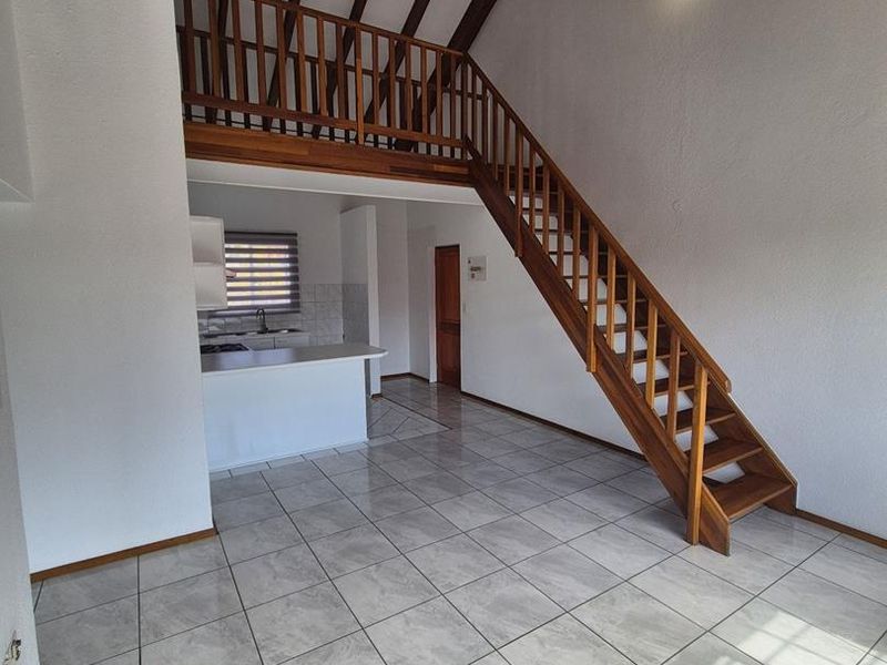 Spacious open-plan living area featuring high vaulted ceilings with exposed timber beams, a wooden staircase leading to a mezzanine level, and tiled flooring throughout.