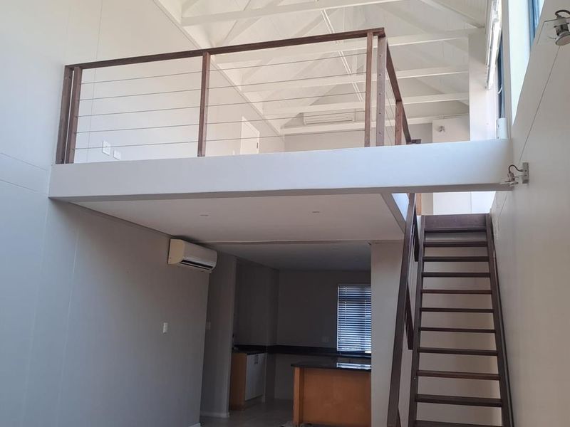 Open-plan living area featuring a mezzanine level, timber staircase, exposed roof trusses, and a functional kitchen space with tiled flooring throughout.