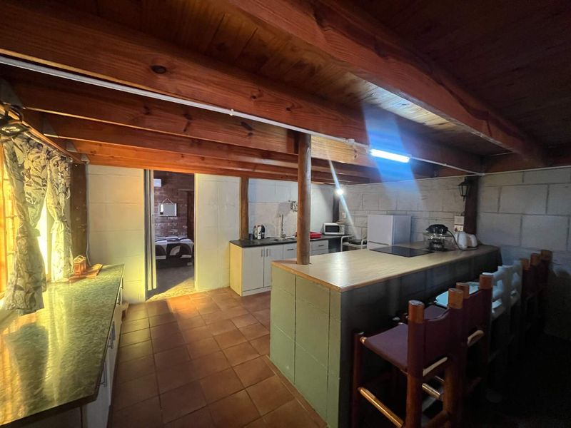 Open-plan kitchen and living area featuring exposed timber ceiling beams, blockwork walls, tiled flooring, and a functional breakfast bar layout.