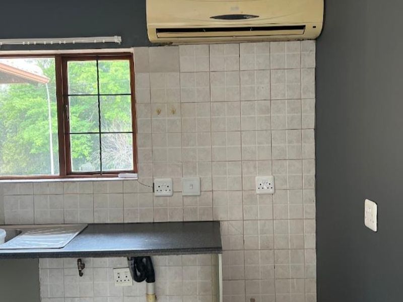 A compact kitchen area featuring a dark countertop, stainless steel sink, tiled splashback, and a wall-mounted air conditioning unit.