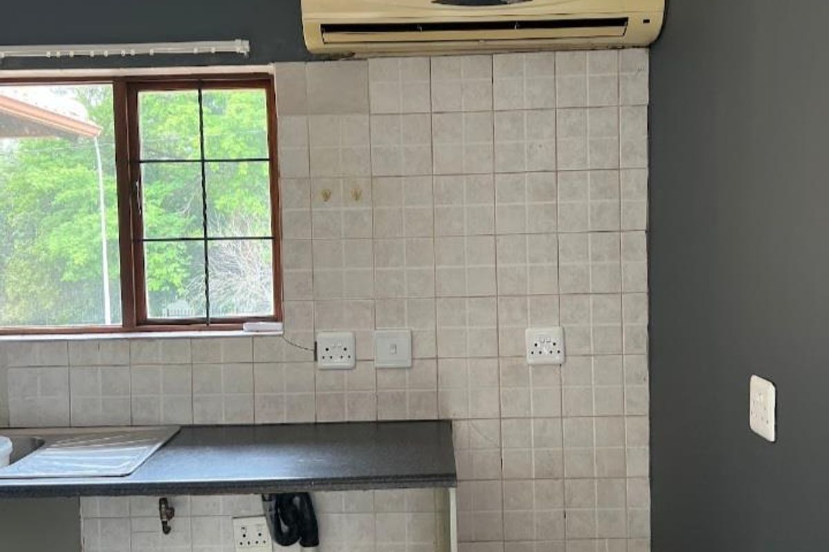 A compact kitchen area featuring a dark countertop, stainless steel sink, tiled splashback, and a wall-mounted air conditioning unit.