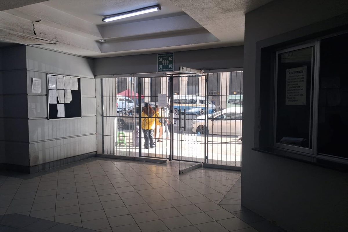 A functional building entrance lobby featuring tiled flooring, a security gate, and a reception window, showing signs of wear on the ceiling.