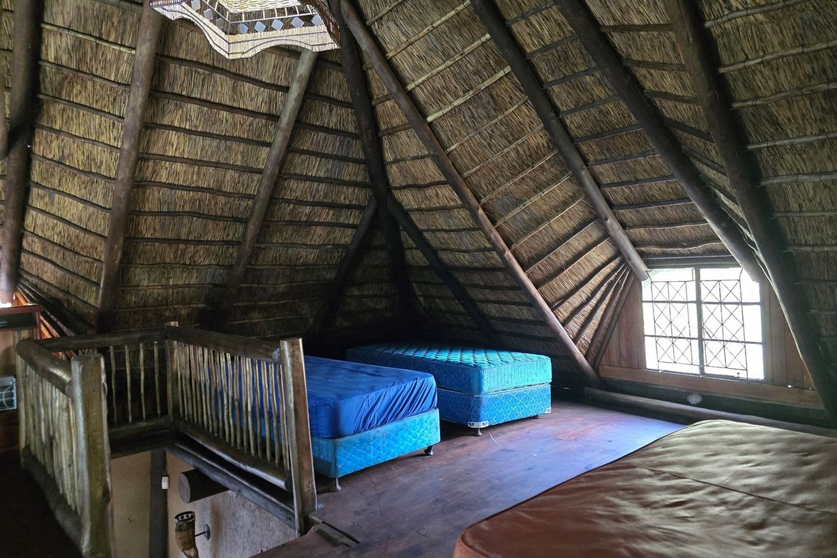 A rustic loft-style bedroom featuring a high vaulted thatch ceiling with exposed timber rafters, timber flooring, and a leaded glass window.