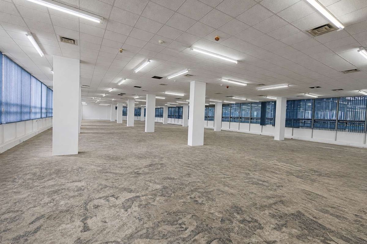 A large, open-plan office space featuring a suspended ceiling, fluorescent lighting, structural columns, and extensive window frontage with vertical blinds.