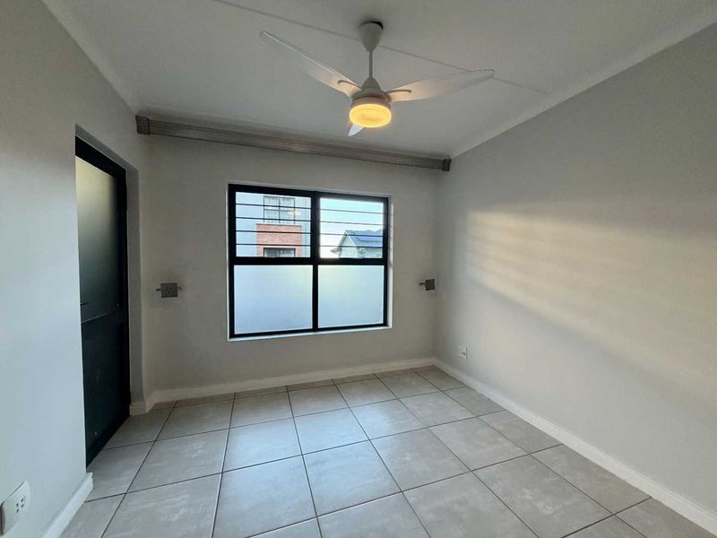 A clean, empty bedroom featuring neutral tiled flooring, a ceiling fan, and a window fitted with security bars.