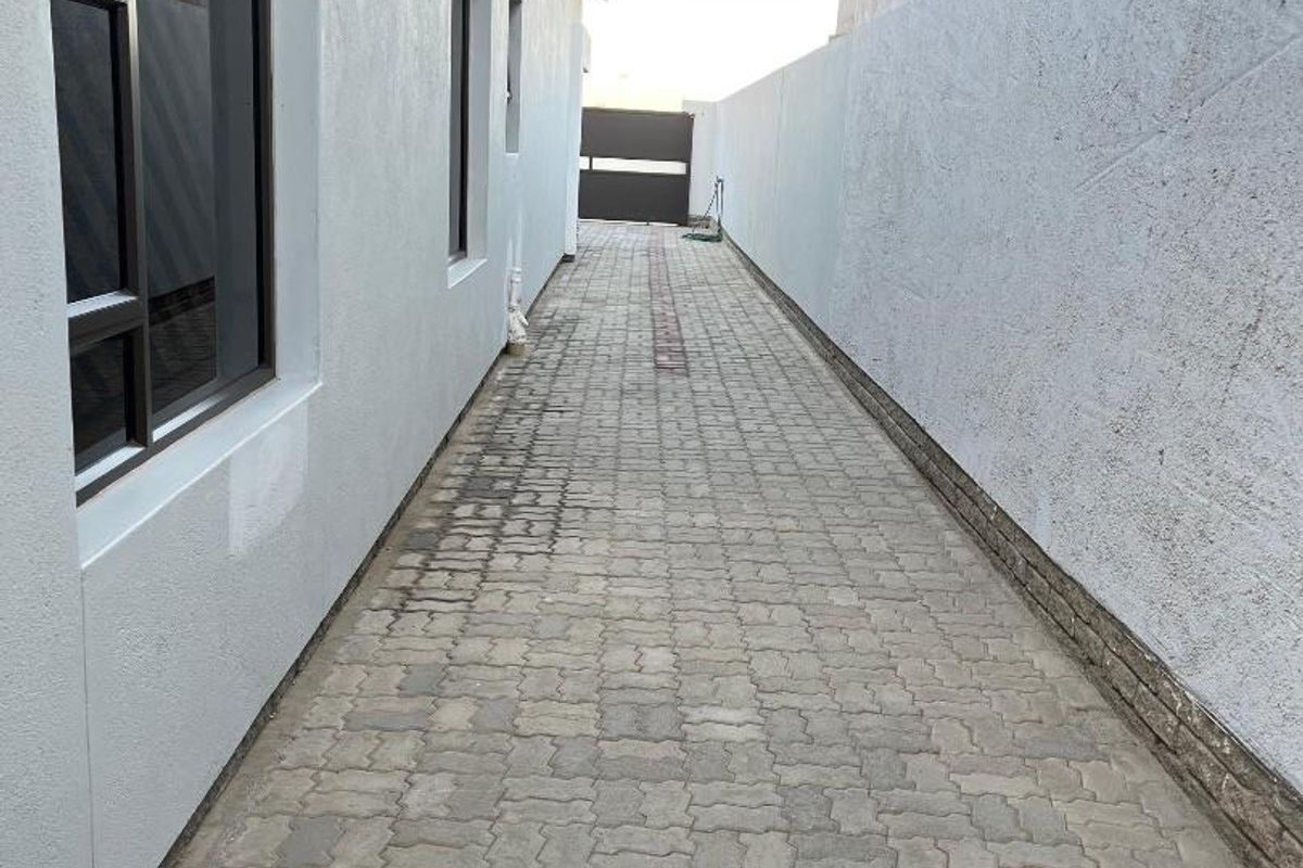A narrow paved side passage or exterior walkway featuring a boundary wall and aluminium window frames along the side of the house.