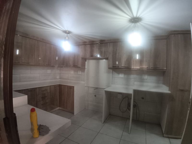 A modern, empty kitchen featuring wood-finish cabinetry, stone countertops, tiled backsplash, and dedicated plumbing connections for appliances.