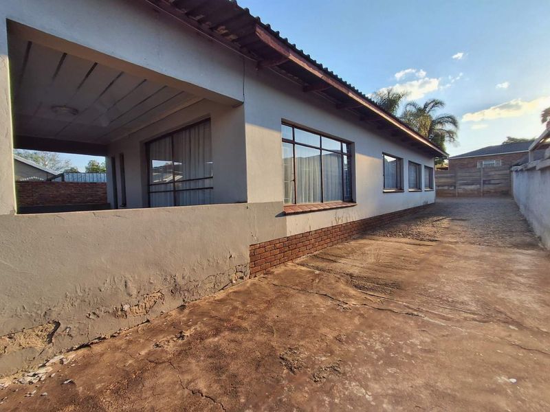 Rear exterior view of the house featuring a covered patio, concrete driveway, and perimeter wall, showing signs of wear on the exterior plaster and driveway.