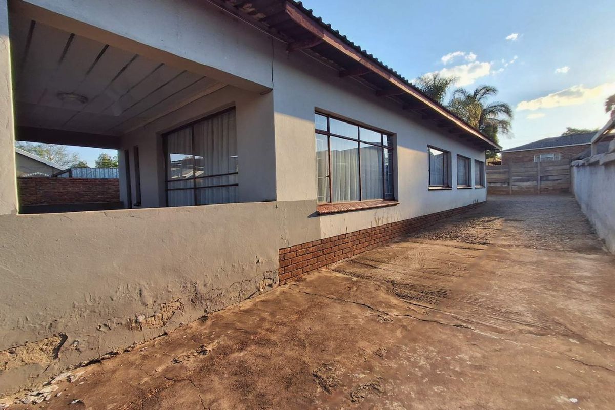 Rear exterior view of the house featuring a covered patio, concrete driveway, and perimeter wall, showing signs of wear on the exterior plaster and driveway.