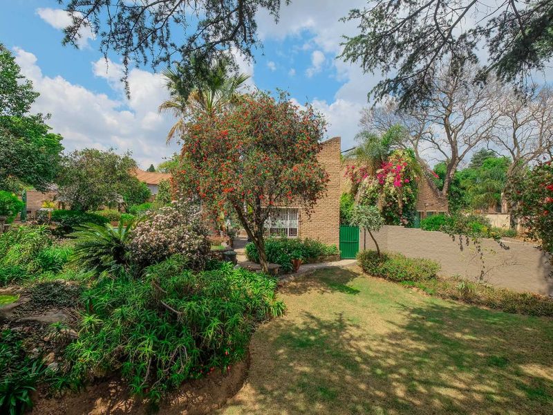 A well-maintained rear garden featuring established trees, landscaped beds, a brick facade building, and a paved pathway leading to a secure gate.