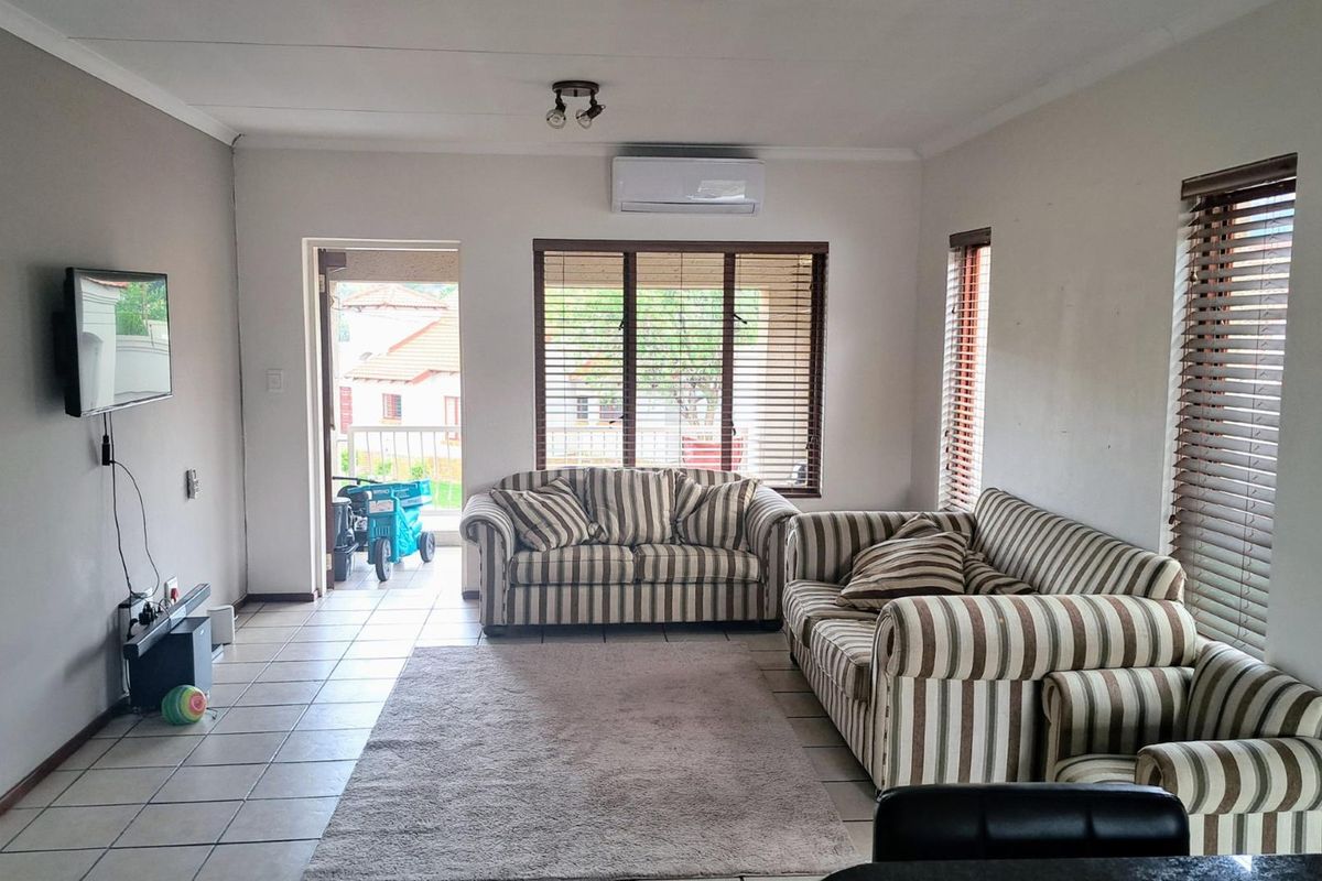 A bright living room featuring tiled flooring, a wall-mounted air conditioning unit, and large windows with fitted blinds, leading out to a patio area.