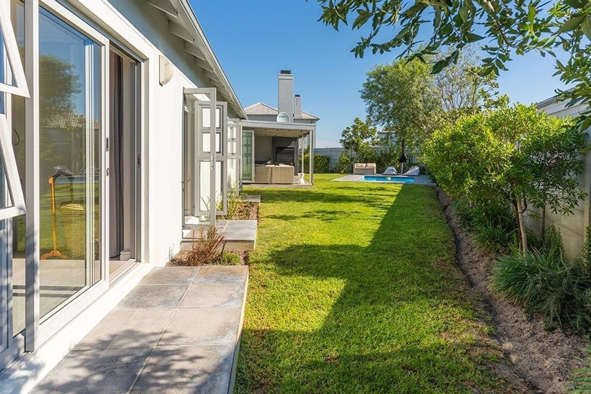 A well-maintained rear garden featuring a manicured lawn, paved patio area, swimming pool, and a covered outdoor entertainment space with a built-in fireplace.