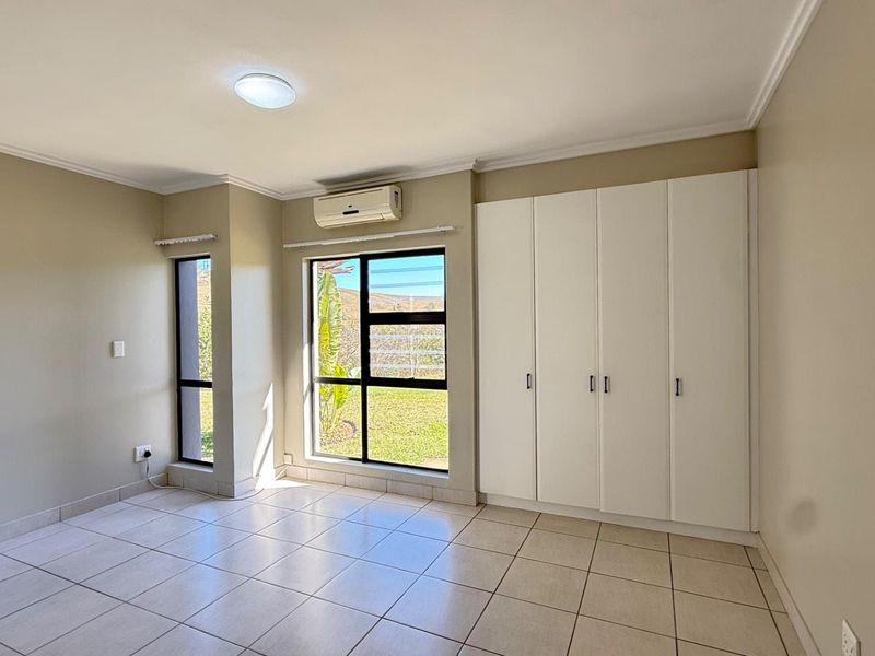 A bright, empty bedroom featuring tiled flooring, built-in cupboards, and an air conditioning unit, with large windows providing natural light.