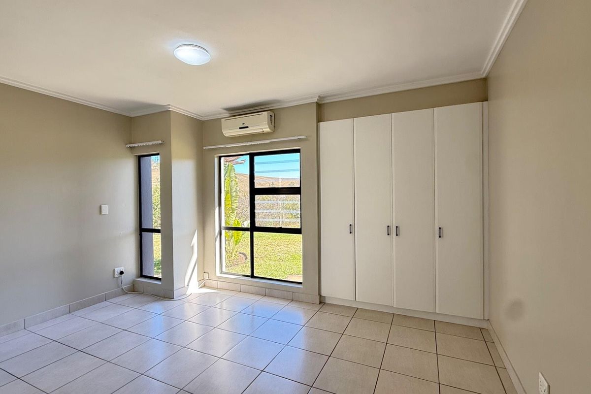A bright, empty bedroom featuring tiled flooring, built-in cupboards, and an air conditioning unit, with large windows providing natural light.