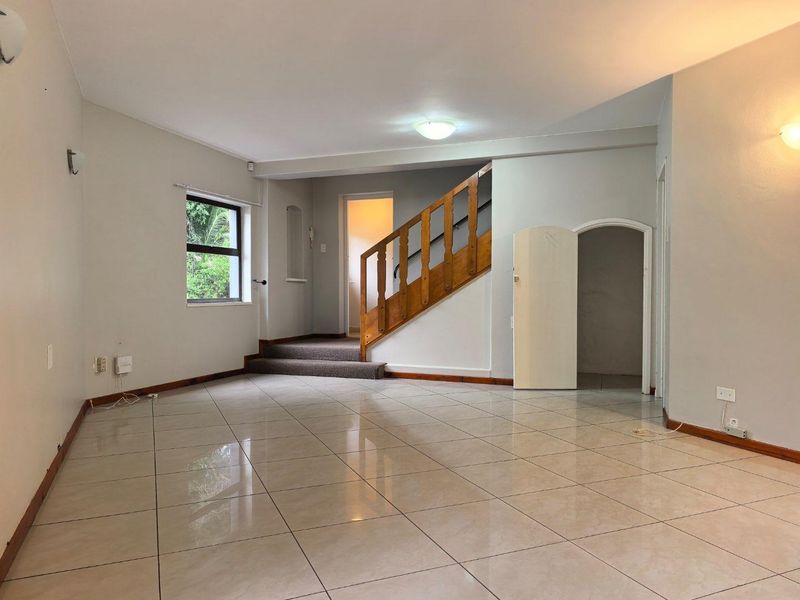 Spacious, empty living area featuring neutral tiled flooring, a wooden staircase, and an arched doorway leading to an adjacent room.
