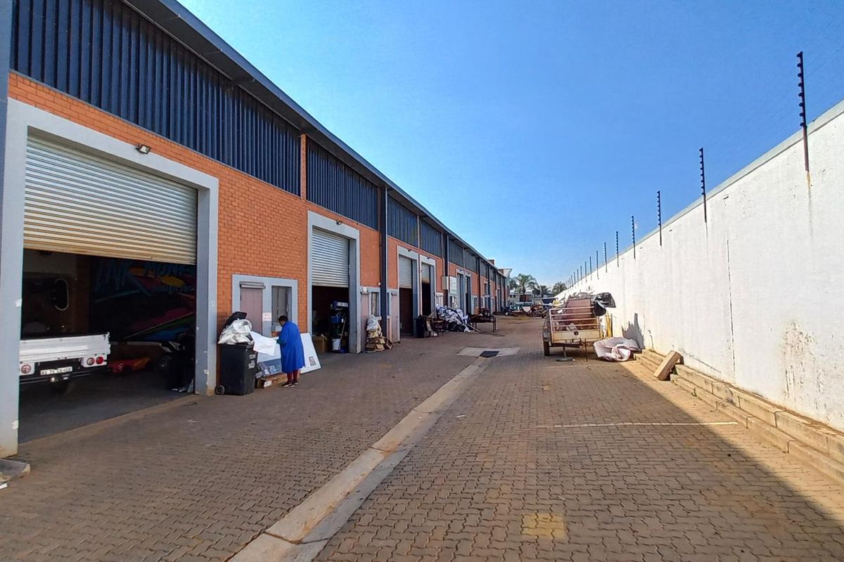 Exterior view of an industrial complex featuring multiple roller shutter doors, brick facade, and a paved driveway enclosed by a high perimeter wall.