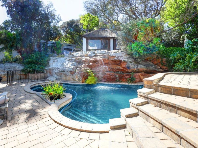 A landscaped pool area featuring a natural rock waterfall, paved patio surround, stone steps, and a charming thatched gazebo in the background.