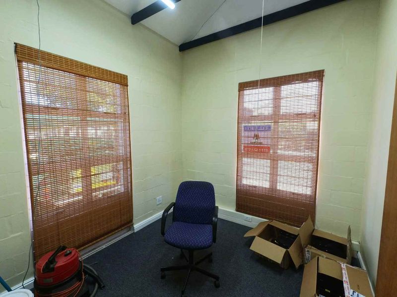A functional office space featuring exposed ceiling beams, carpeted flooring, and natural light provided by two windows with bamboo blinds.