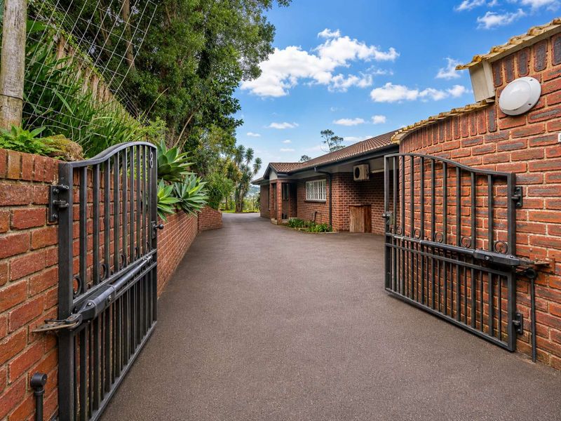 A well-maintained brick townhouse exterior featuring a paved driveway, secure metal entrance gate, and established garden borders.