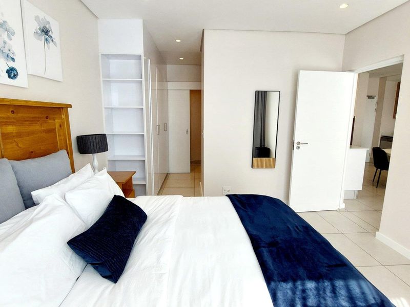 A well-maintained bedroom featuring neutral walls, tiled flooring, built-in storage cupboards, and recessed ceiling lighting, with an adjoining doorway leading to another living space.