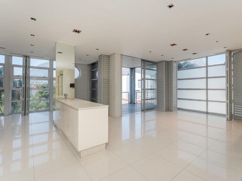 Spacious open-plan living area featuring polished tiled floors, recessed lighting, a central kitchen island, and large floor-to-ceiling windows with integrated folding shutters.