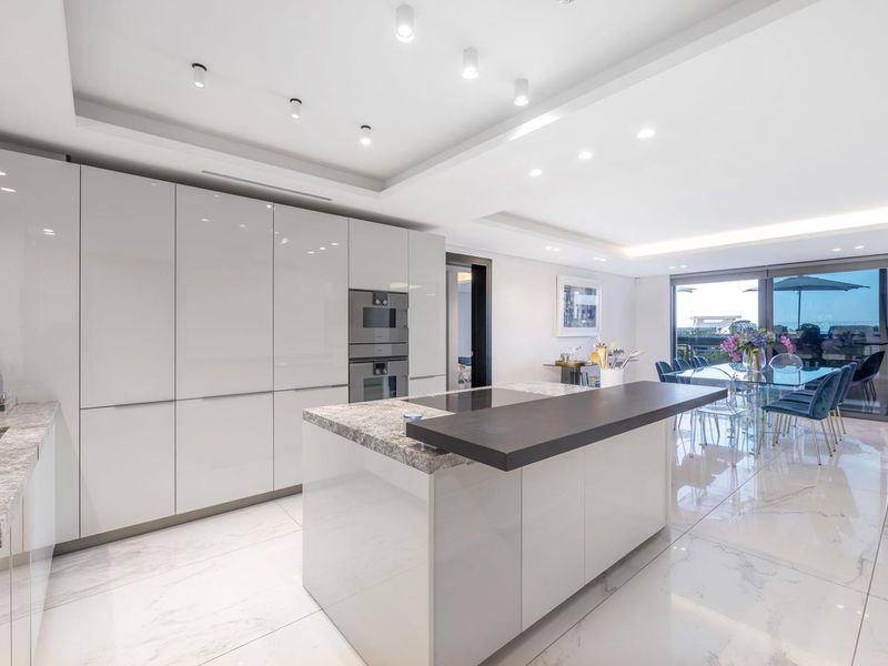 Modern open-plan kitchen and dining area featuring high-gloss cabinetry, stone-topped island, integrated appliances, and polished marble-look flooring with seamless access to the balcony.