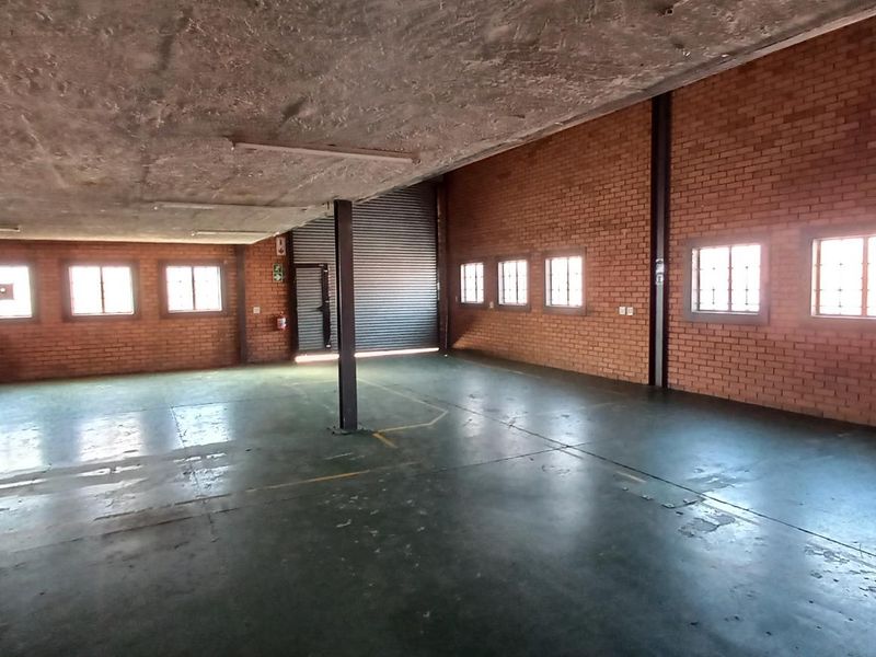 Spacious industrial warehouse area featuring exposed brick walls, concrete ceiling, multiple windows for natural light, and a large roller shutter door.