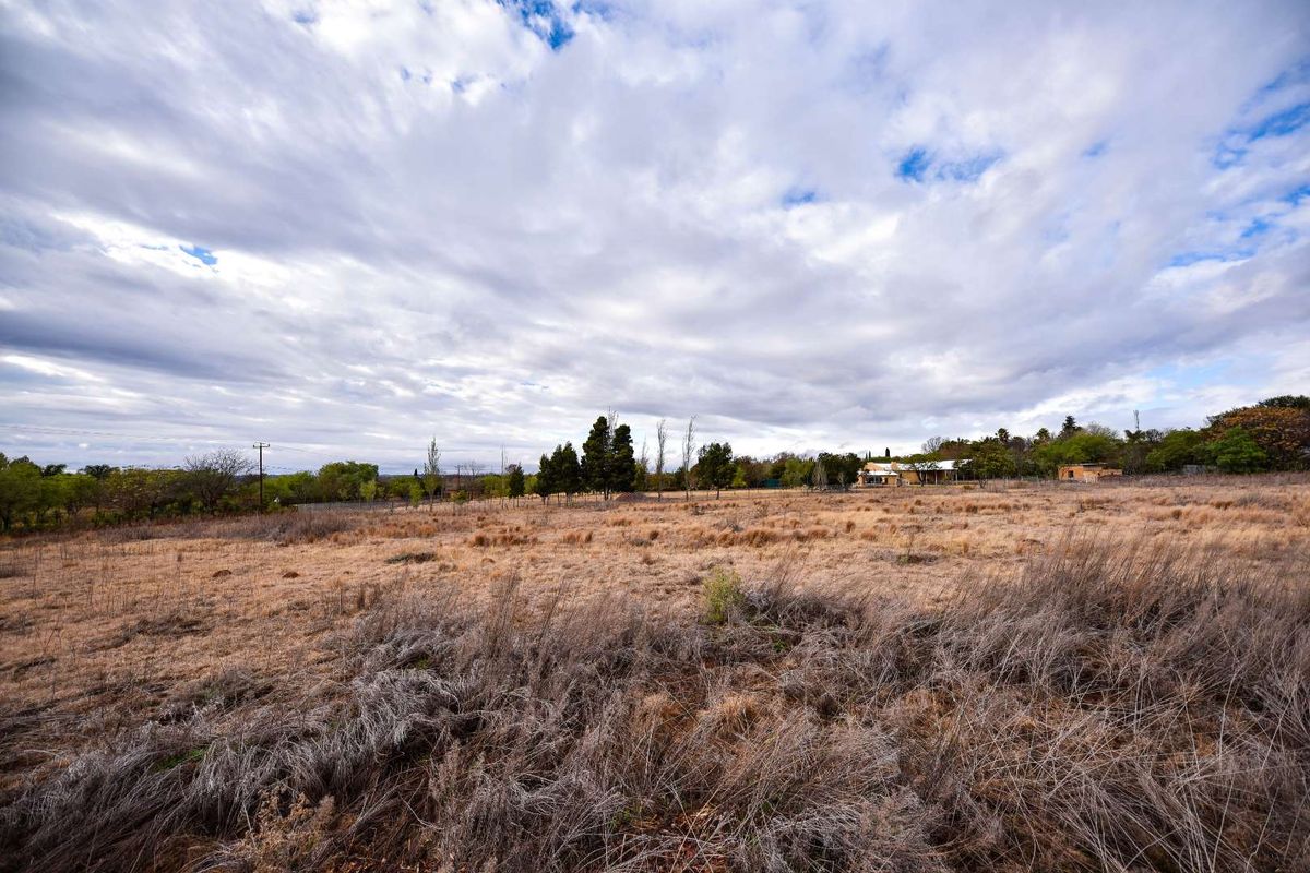 A wide-angle view of a large, undeveloped residential plot featuring dry grass, perimeter fencing, and mature trees under a cloudy sky.