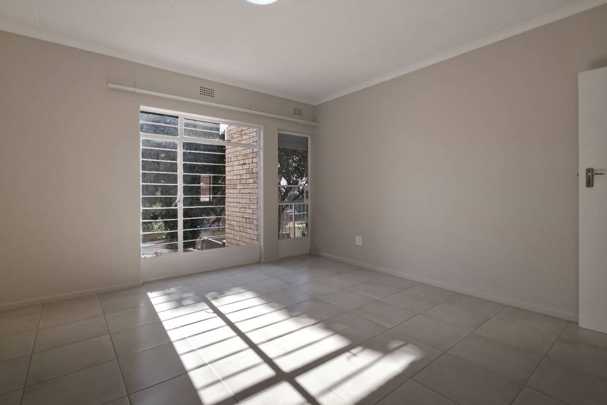 A bright, empty bedroom featuring neutral tiled flooring, large windows with security bars, and a door providing direct access to the exterior.