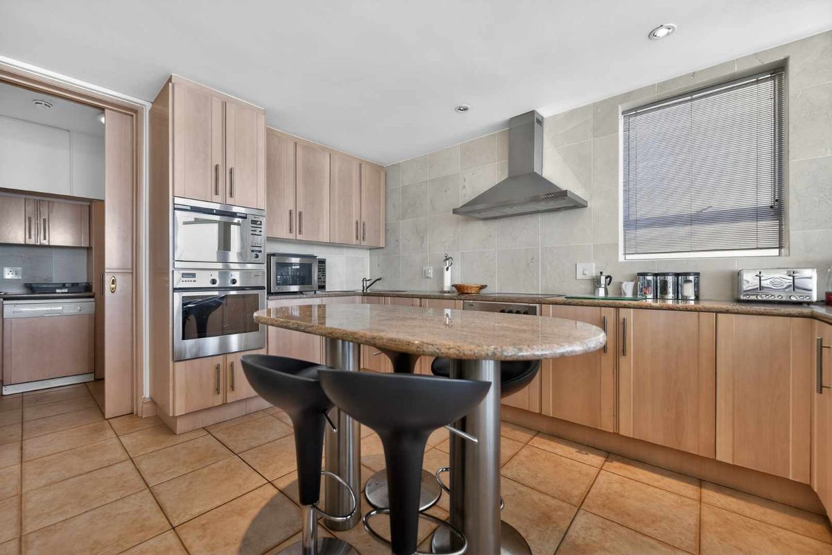 A functional kitchen featuring light wood cabinetry, granite countertops, built-in stainless steel appliances, and a breakfast bar, with an adjacent scullery area.