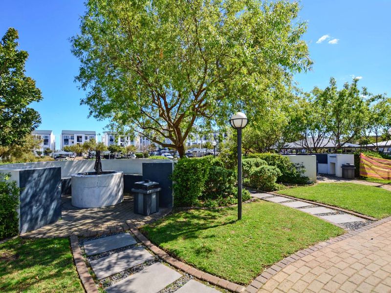A well-maintained communal garden area featuring paved walkways, established trees, and a built-in braai facility for outdoor entertainment.