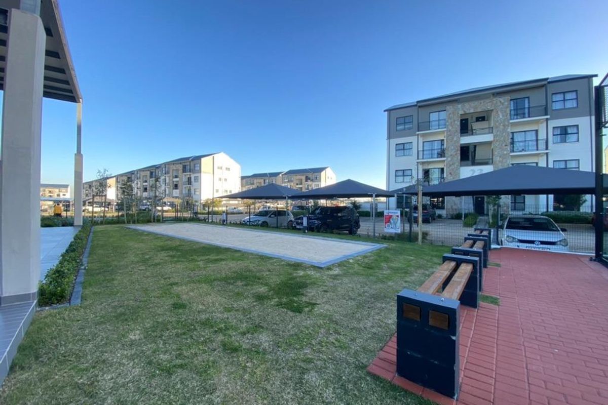 Communal outdoor area featuring a sand pit, paved walkways, and shaded parking structures within a modern apartment complex.