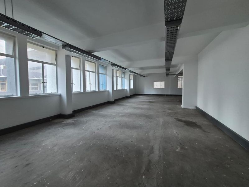 A spacious, vacant open-plan office area featuring large windows, exposed ceiling lighting tracks, and unfinished concrete flooring, ready for custom fit-out.