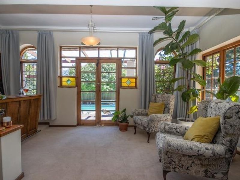 A living room featuring a built-in timber bar, carpet flooring, and timber-framed french doors with stained glass accents, providing direct access to the pool area.