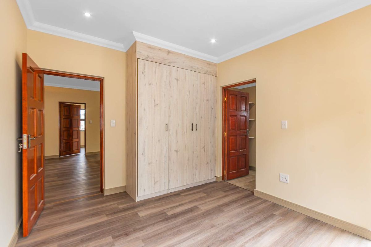 A clean, empty bedroom featuring modern light-wood built-in wardrobes, laminate flooring, and recessed downlights, with access to adjacent rooms through timber doors.