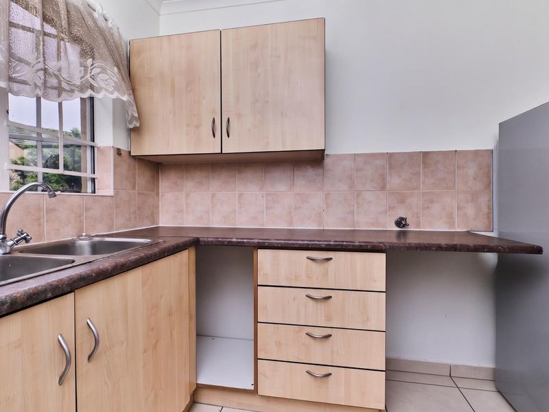 A functional kitchen featuring light wood-tone cabinetry, laminate countertops, a stainless steel double sink, and tiled splashbacks with neutral floor tiling.