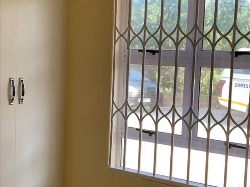 A clean, empty bedroom featuring built-in cupboards, tiled flooring, and a window fitted with a security gate.