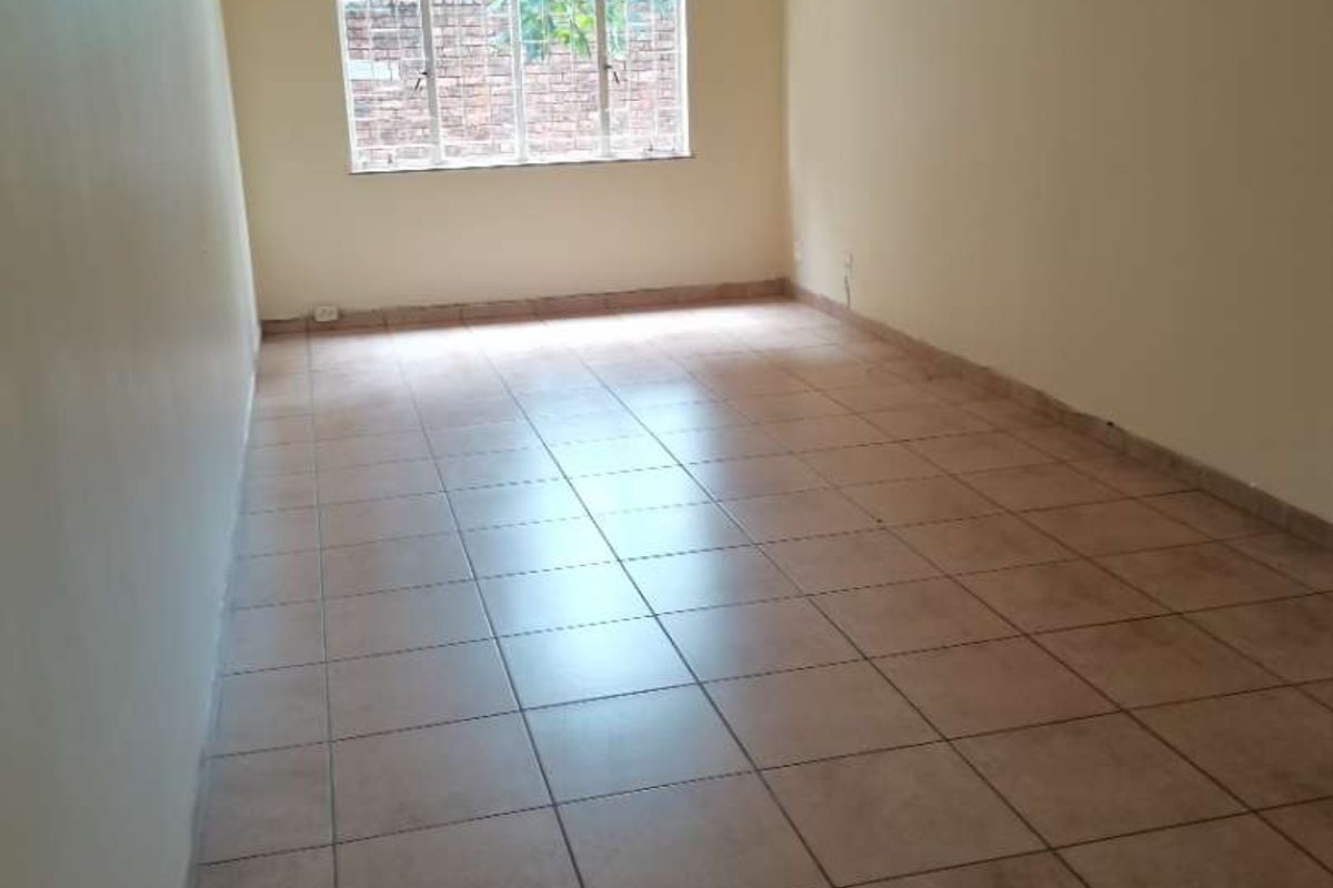 A spacious, empty living area featuring neutral tiled flooring, a ceiling fan, and a large window providing natural light.