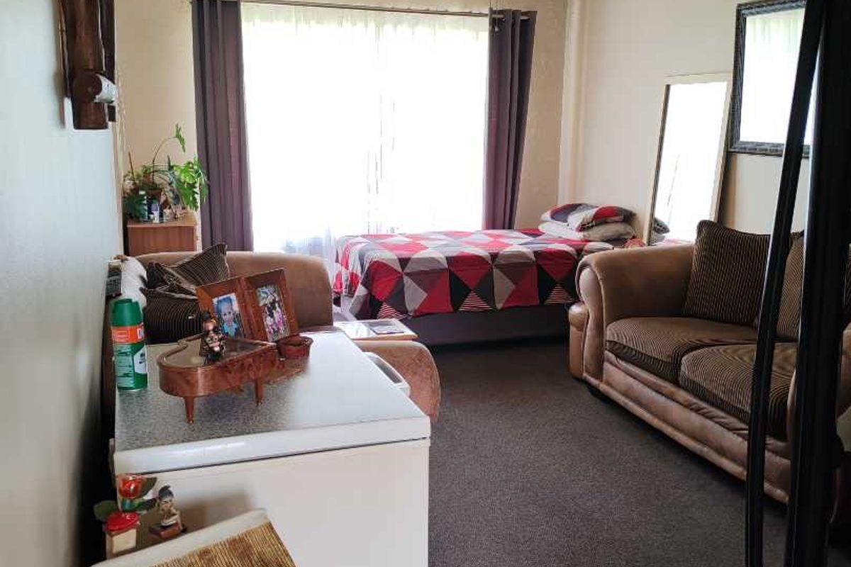 A multi-purpose living and sleeping area featuring a ceiling fan, large window with curtains, and carpeted flooring.