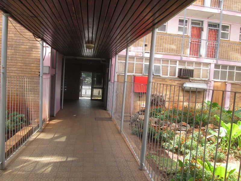 A covered walkway leading to the apartment entrance, featuring brick facade walls, metal security railings, and tiled flooring within a communal complex.