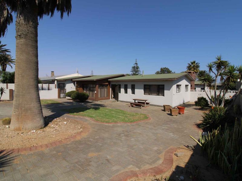 Rear view of the property featuring a paved courtyard, landscaped garden areas, and a single-story house with a metal roof.