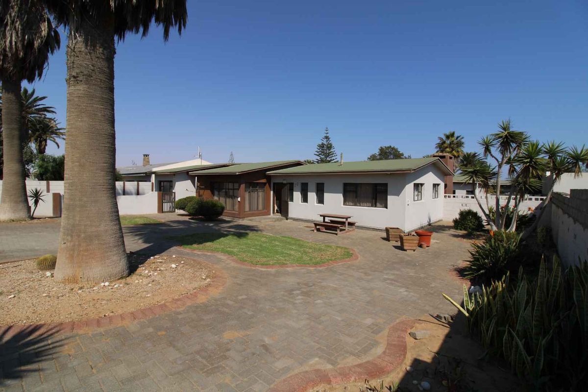 Rear view of the property featuring a paved courtyard, landscaped garden areas, and a single-story house with a metal roof.