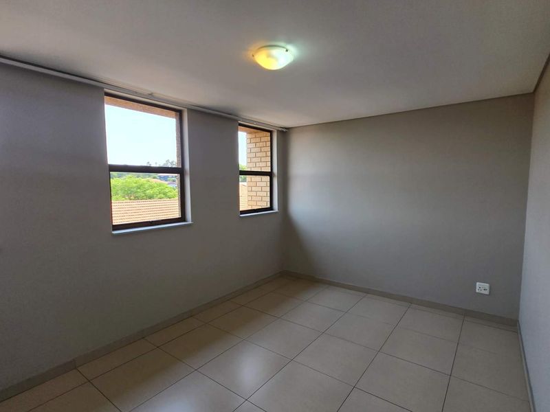 A bright, empty bedroom featuring neutral tiled flooring, clean walls, and large windows providing natural light and a pleasant exterior view.