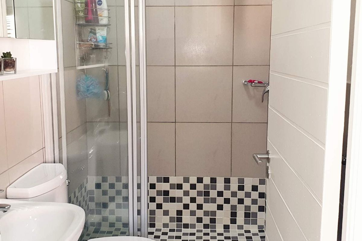A compact bathroom featuring a glass-enclosed shower, tiled walls with a decorative mosaic accent, a pedestal basin, and a modern toilet.
