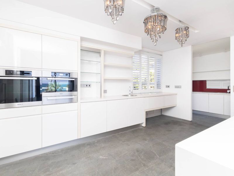 Modern, minimalist kitchen featuring high-gloss white cabinetry, built-in Miele appliances, concrete flooring, and elegant crystal chandeliers, with an adjacent scullery area.
